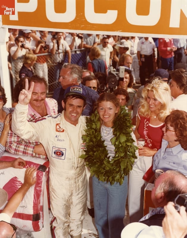 Al Unser, Winner of the Pocono 500 Race in 1978, with Jeanetta Holder's Quilt at his Side Man in white jumpsuit with arm around woman with large laurel wreath around her neck stand surrounded by other people, folded quilt to one side