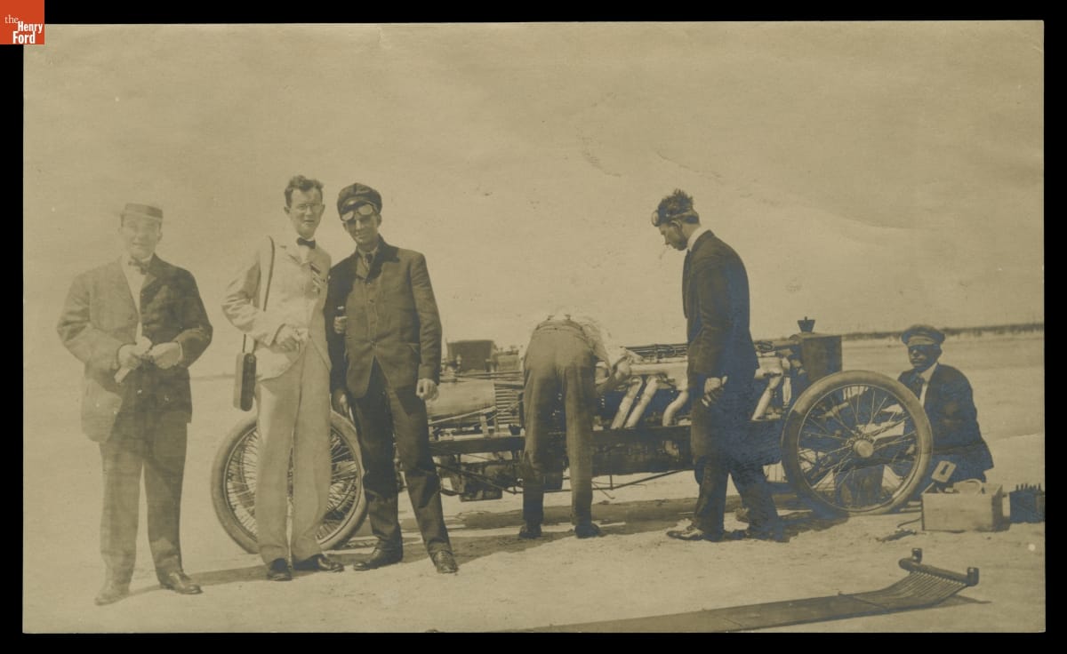 Black-and-white photo of three men standing near and three men working on early open race car on a beach