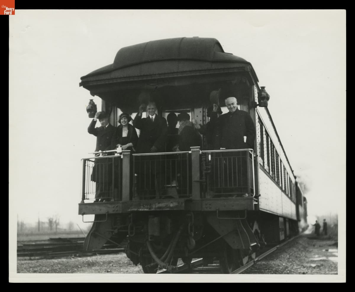 Several people stand on the back platform of a railcar, some waving