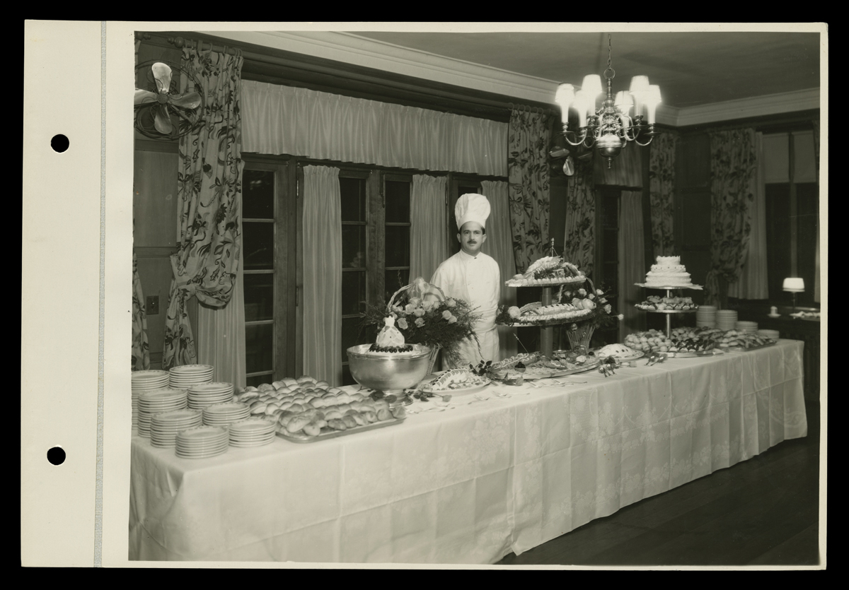 Man in chef's outfit and hat stands behind long buffet table filled with plates and displays of food 