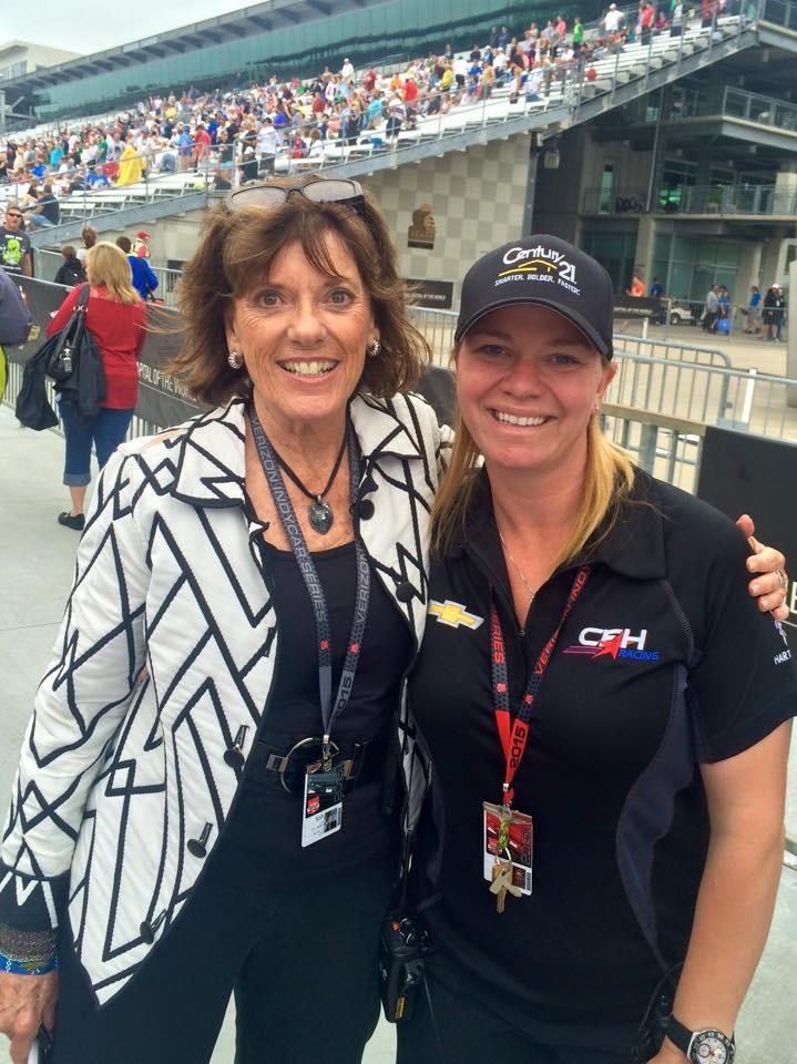 Two women wearing lanyards pose with arms around each other, with a grandstand full of people in the background