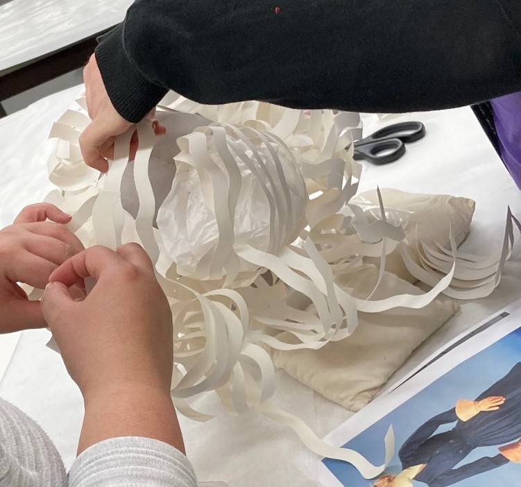Adding a ball of tissue paper for volume and extra support An arm extends, holding up a complex white cut paper shape, while two hands in lower left also hold the shape