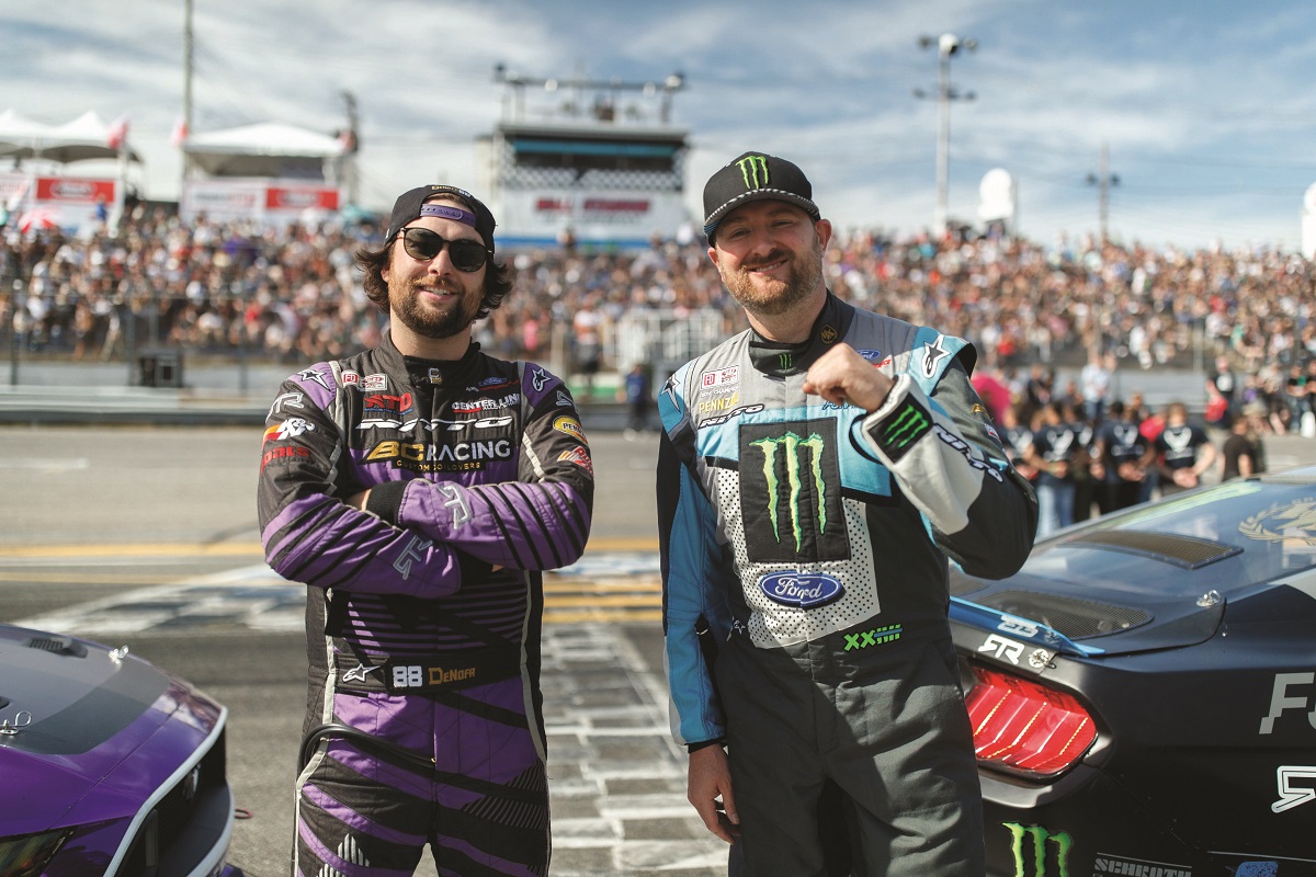 Vaughn Gittin, Jr., and Chelsea DeNofa at Formula Drift New Jersey 2019 Two men in racing jumpsuits and baseball caps stand by cars in front of a track and spectators in grandstands