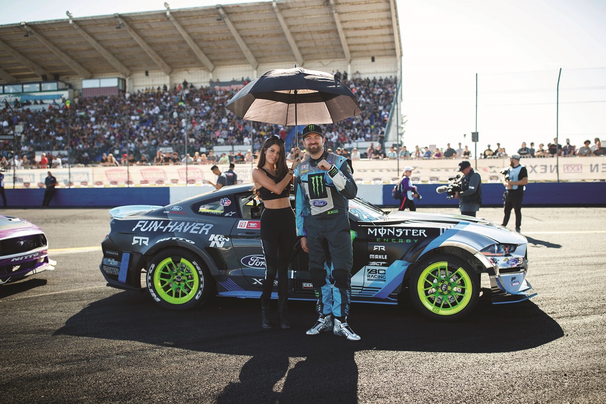 Vaughn Gittin, Jr., and Christen Dye Woman holding umbrella stands with man in racing jumpsuit in front of racecar on track with spectators in grandstands behind them