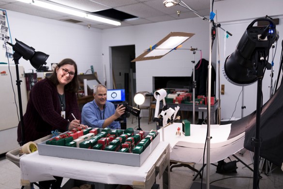Man points camera at photo set and woman works at cart filled with small green and red boxes