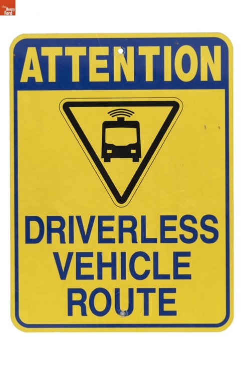 Yellow sign with text "ATTENTION DRIVERLESS VEHICLE ROUTE" and icon of a van in upside-down triangle
